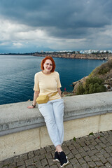 Fototapeta premium Young smiling redhead Caucasian woman posing for photographer while sitting on fence of observation point overlooking sea in Antalya.