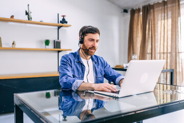 Mature bearded man in headphones working at the table with laptop at office.