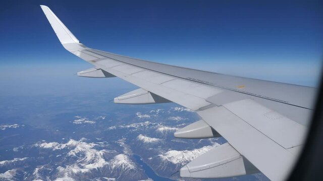View from passenger cabin window of wing of aircraft flying over Austrian Alps on sunny day against background of blue sky and landscape with snow-capped rocky mountain ranges below. Travel concept