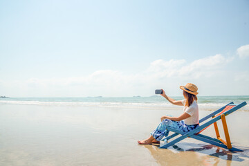 Asian woman resting on sunbed on the beach. Happy girl sit on beach chair using smartphone selfie...