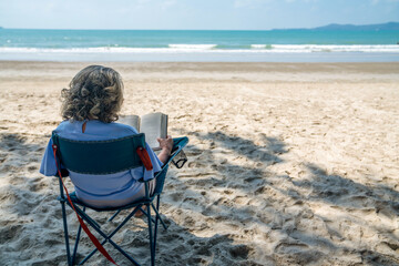Happy Asian senior woman sitting on outdoor chair on the beach with reading a book. Healthy...
