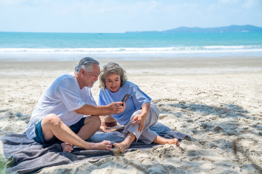 Happy Asian family on beach vacation. Senior couple sit on the beach using smartphone for online shopping or video call. Healthy elderly husband and wife enjoy outdoor lifestyle together in summer. - Powered by Adobe
