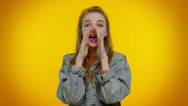Hey you. Teenager student girl 20s years old in denim jacket scream with hand near mouth waving and greeting with hand as notices someone shouting hello. Young woman posing on yellow studio background