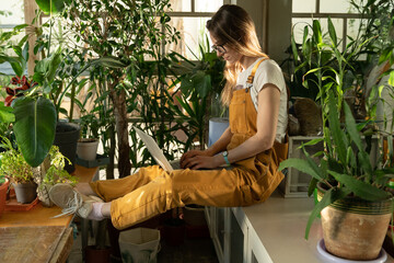 Young girl in jumpsuit browse on laptop in green house. Female gardener use computer for education, online orders, communication with clients. Young entrepreneur or freelancer working from home office