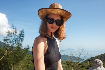 close up portrait of young blonde woman traveler in hat and sunglasses at sunny summer day on mountains and sea background