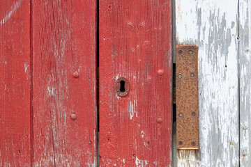A closeup of a bright red vintage metal keyholder in a textured red wooden door. The exterior of the old woodshed has worn and wear patterns with some scuff marks.The door is on a white wood building.