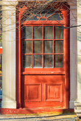 Obraz premium An exterior wide vintage vibrant red wooden decorative panel door with multiple small glass panes in a white building. There's an arched window glass over the door. The sun is shining on the door. 