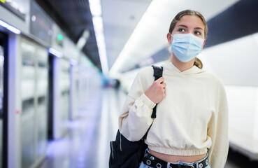 Fototapeta na wymiar Young woman in face mask waiting for train on platform of subway underground station