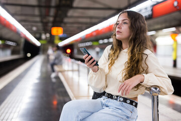 Young attractive girl in jeans waiting for subway train on a platform, using her smartphone
