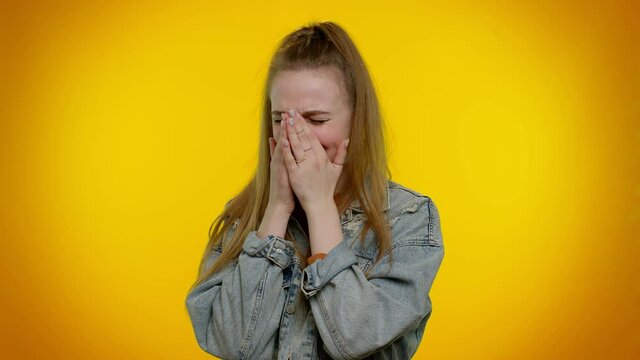Upset disappointed pretty stylish teen girl in denim jacket wipes tears and cries from despair, being sad because of unfair things. Young adult woman. Indoor studio shot isolated on yellow background