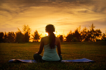 Concept of healthy lifestyle and relaxation-back view of attractive girl in sportswear with headset sitting in lotus position while listening the music outdoor on sunset