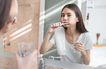 Young Asian woman brushing teeth and looking in the mirror, holding water glass, towel on the shoulder on bathroom, Concept oral hygiene and health care. - Powered by Adobe
