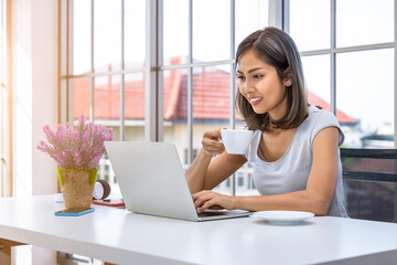 Portrait of Asian woman freelancer working at home, sitting at desk table in living room, drinking coffee