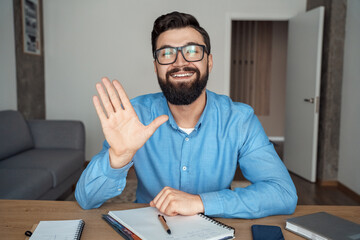 Smiling millennial caucasian man at office desk looking at camera waving hand