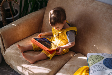 A small child watches cartoons on a tablet while sitting on the couch at home alone.