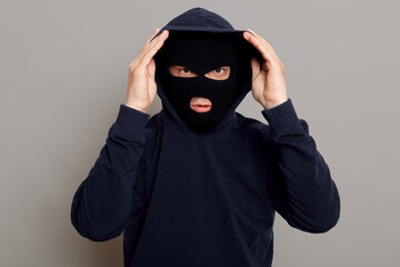 Concentrated young criminal man in a bandit mask dressing a hood, going to rob someone, trying to...