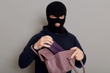 A criminal thief takes a wallet from a stolen bag, dressed in a black hoodie and a robbery mask,...