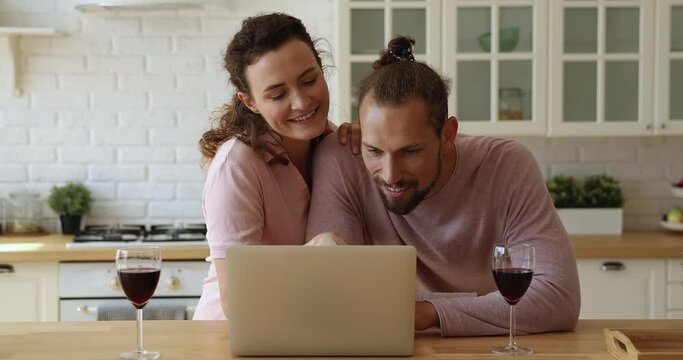 Happy young loving couple using laptop, standing at table with glasses with red wine, planning wedding celebration, honeymoon trip or vacation travel web surfing information online together.