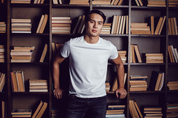 Asian man in white T-shirt stands against the background of bookshelves. Mockup.
