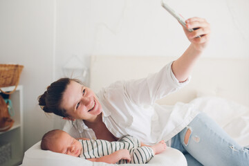 Cute caucasian mom with a newborn baby. A mother in a white shirt takes a selfie on the phone, the baby lies in a cocoon in  cradle for newborns, light toning.  social networks and video communication