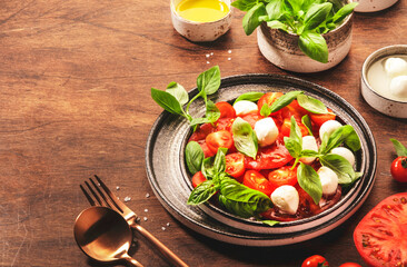 Summer italian salad Caprese with red tomatoes and mozzarella cheese with green basil leaves and olive oil dressing. Top view, wooden table