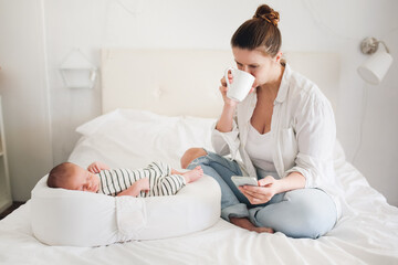 Cute caucasian mom with a newborn baby. Mother with phone and coffee while baby is sleeping,...