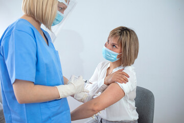 Female doctor holding syringe making covid 19 vaccination injection dose in shoulder of female patient wearing mask. Flu influenza vaccine clinical trials concept, corona virus treatment