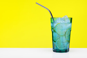 Refreshing drink with ice, lemon and lime wedges. Large blue glass with a drinking straw, yellow background.