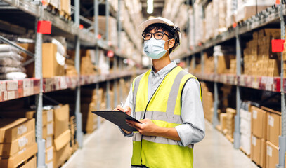 Portrait of asian engineer man in helmet in quarantine for coronavirus wearing protective mask order details checking goods and supplies on shelves with goods background in warehouse.logistic
