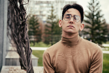 Portrait of a handsome young man with a stylish haircut posing in a brown turtleneck. Strong look and face. Shooting outdoors. Cute student with glasses.
