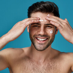Happy brunette guy smiling at camera while applying white cream on his face, posing isolated over...