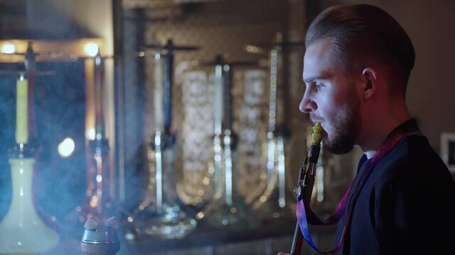 A young guy lights a hookah behind the bar and blows smoke rings