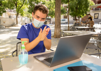 Young man with protective face mask and hand sanitizer outdoors working remotely on computer
