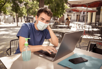 Young man with protective face mask and hand sanitizer outdoors working remotely on computer