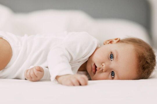 A blue-eyed baby lies in a snow-white bed under a blanket, laughs and indulges
