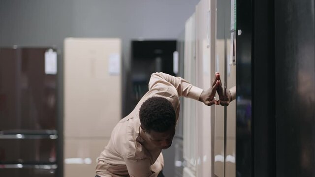 young black man is viewing fridge with freezer in home appliance store, examining exhibition sample, opening doors and looking inside