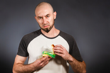 Environmental pollution and food industry. Portrait of a bald man with a beard, who in bewilderment reveals a hamburger stuffed with garbage. Grey background