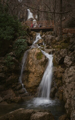 Fototapeta premium The waterfall is a gloomy forest, The water falls in a large stream from the mountains. Vertical natural autumn background. Tourist route to the waterfall. Unrecognizable people behind in the distance