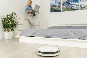 happy woman using laptop while robotic vacuum cleaner washing in living room