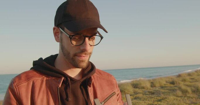 Giovane ragazzo con occhiali e cappello pilota il drone in spiaggia vicino al mare
