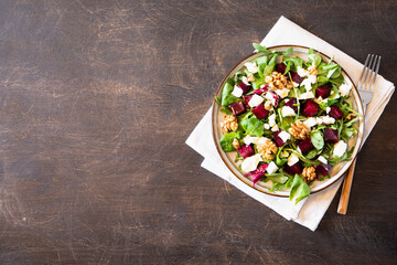 Beet or beetroot salad with fresh arugula, soft cheese and walnuts on plate, dressing and spices on dark wooden background, copy space, top view