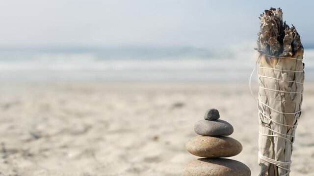 Dried white sage smoke, smudge stick burning in soft focus with bokeh, aroma smudging close up. Rock balancing on ocean beach, stones stacking by sea water waves. Zen pyramid of pebbles on sandy shore