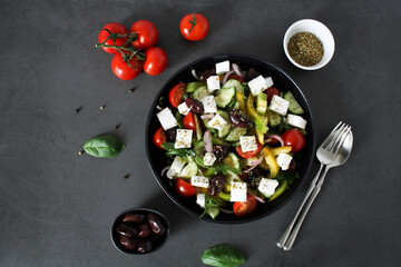 Greek salad - vegetable salad of fresh cherry tomatoes, bell peppers, feta cheese, purple onions and olives in a black plate with a fork on a dark background