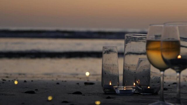 Candle flame lights in glass, romantic beach date by California ocean waves, summer sea water. Candlelight. Wineglass, glass with white wine on sand. Cozy twilight lounge. Seamless looped cinemagraph