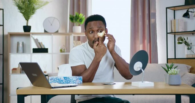 Handsome positive young african american sitting in front of small mirror at home and putting golden patchi under eyes, face care concept