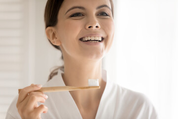 Happy young woman satisfied with wooden bamboo toothbrush, bio whitening toothpaste, holding brush, smiling at mirror reflection, laughing. Sustainable eco friendly production concept. Head shot
