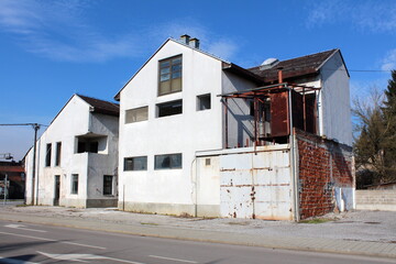 Large abandoned suburban family house with cracked dilapidated white facade and rusted sliding metal garage doors next to paved street at old part of town surrounded with tall trees and blue sky