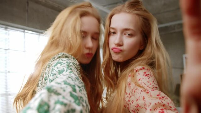 Two young sisters Twins fool around at the camera, laugh, shook, show tongues. Shot of Selfie at home in the living room.