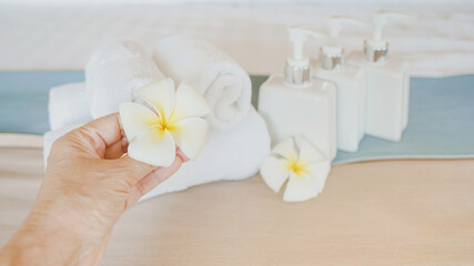 human's hand holding white blossom plumeria flower with blurred bath accessories on bed background for resort , hotel , spa welcome guest concept
