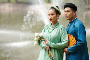 Serious beautiful young Vietnamese just married couple in traditional dresses and headwear standing...